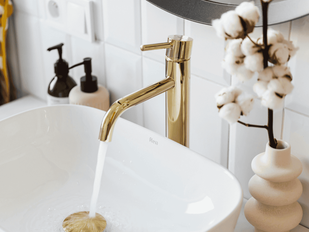 Luxury gold faucet in a modern bathroom with white tile background and decorative cotton branch vase.